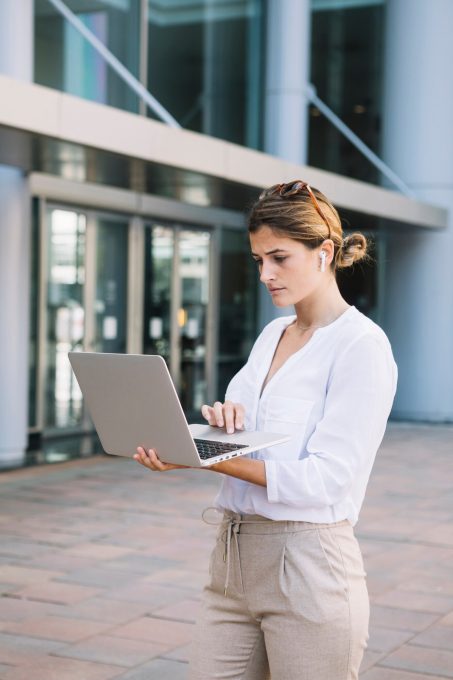 serious-young-businesswoman-standing-outside-her-office-building-using-laptop_23-2147972942