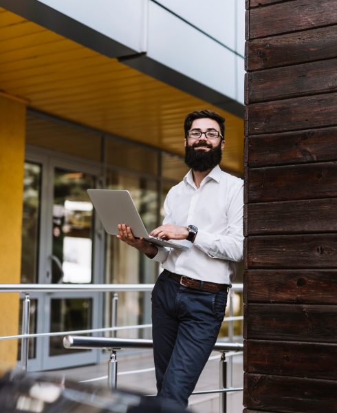 smiling-young-businessman-holding-laptop-hand-leaning-wall_23-2147941454